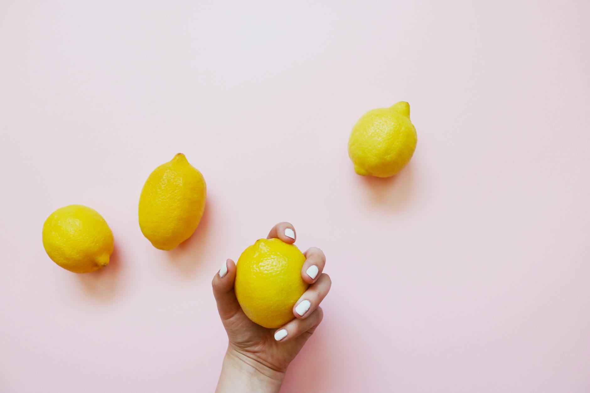 A hand holding a lemon against a soft pink background with additional lemons nearby