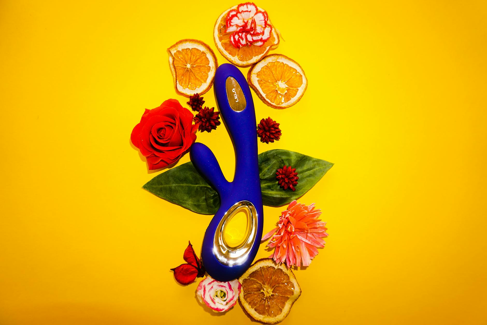Blue lemon vibrator surrounded by fresh flowers and citrus slices on a bright yellow background.